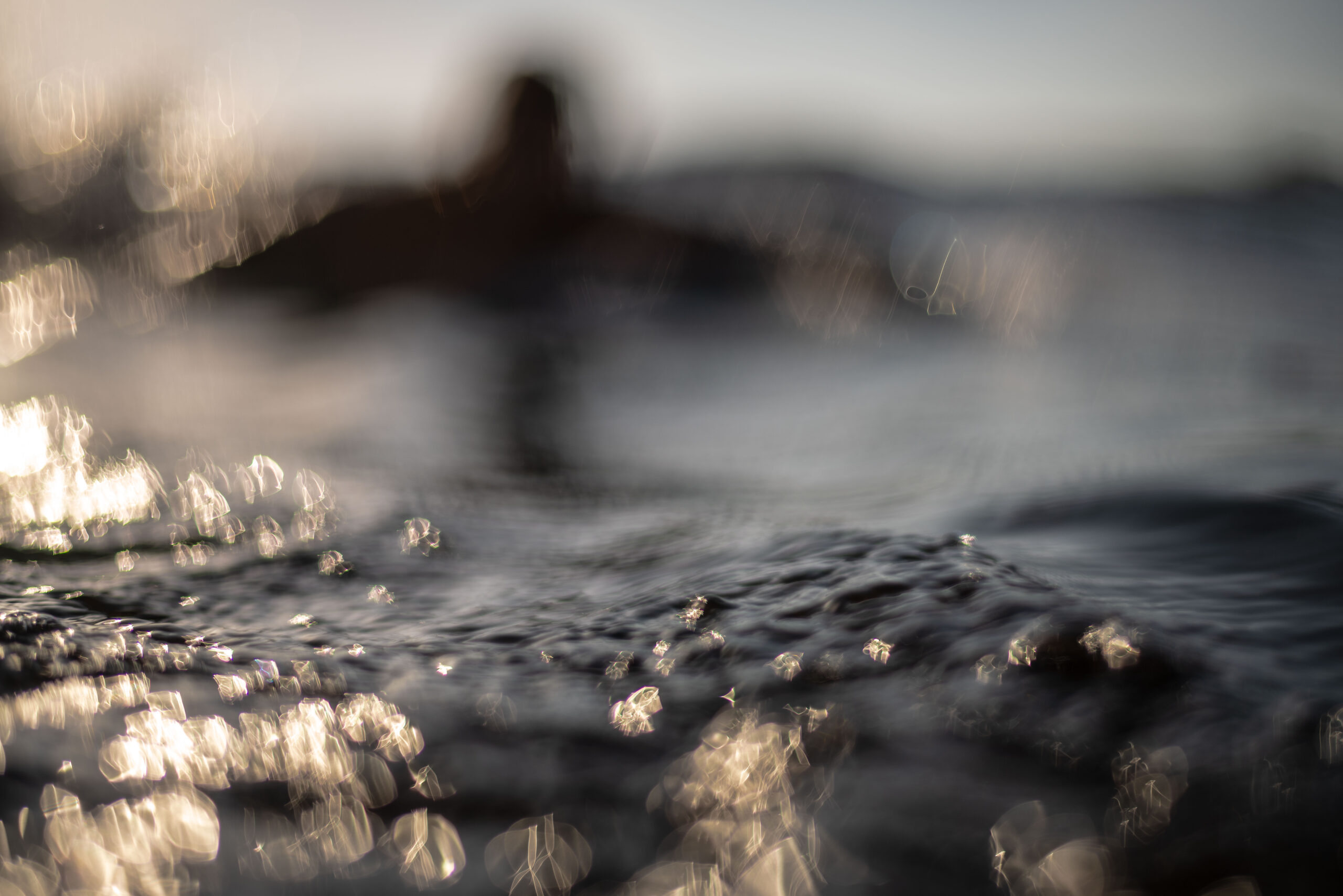 Encinitas surf art, an abstract image of a surfer surfing at Beacons Beach in Encinitas. The photo is focused on ripples of water in the foreground and a silhouette of a surfer paddling for a wave in the background.